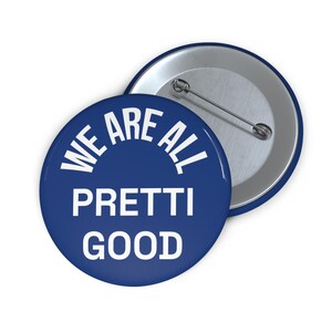May include: Two round blue buttons with the text "WE ARE ALL PRETTI GOOD" in white. One button is face up, and the other is face down, showing the pin back. The buttons have a shiny finish.