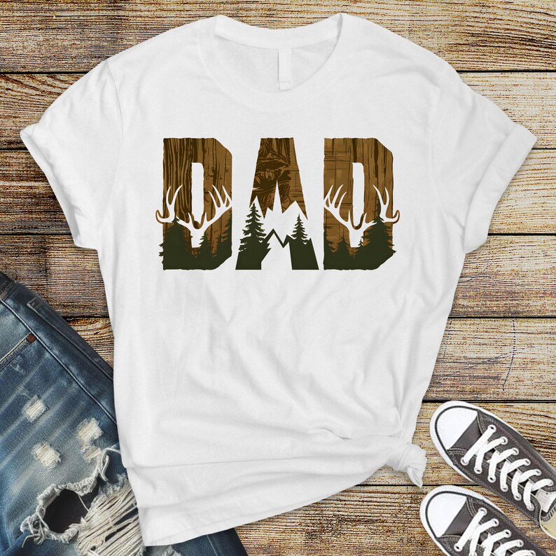 May include: White t-shirt with a brown and green graphic of the word "DAD" with a deer silhouette and a mountain scene.