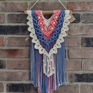 May include: A macrame wall hanging with a blue, pink, and cream color scheme. The hanging features a geometric pattern with wooden beads and a wooden dowel at the top.