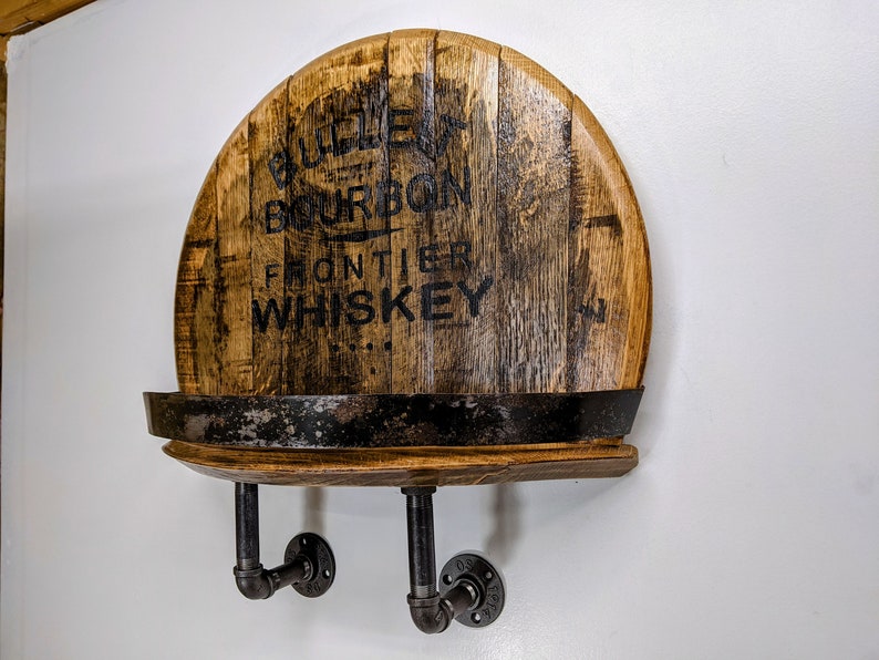 Op de afbeelding: Een houten plank gemaakt van een hergebruikt whiskeyvat met zwarte metalen pijpbeugels. Het vat is gelabeld met "Old Frontier Bourbon Whiskey".