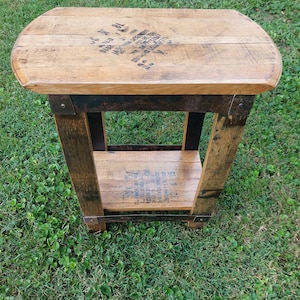 May include: A small wooden table crafted from reclaimed whiskey barrels. The table features a single shelf and is stained a dark brown colour. The tabletop is made from a single piece of wood with a rounded edge. The table is positioned on a patch of green grass.