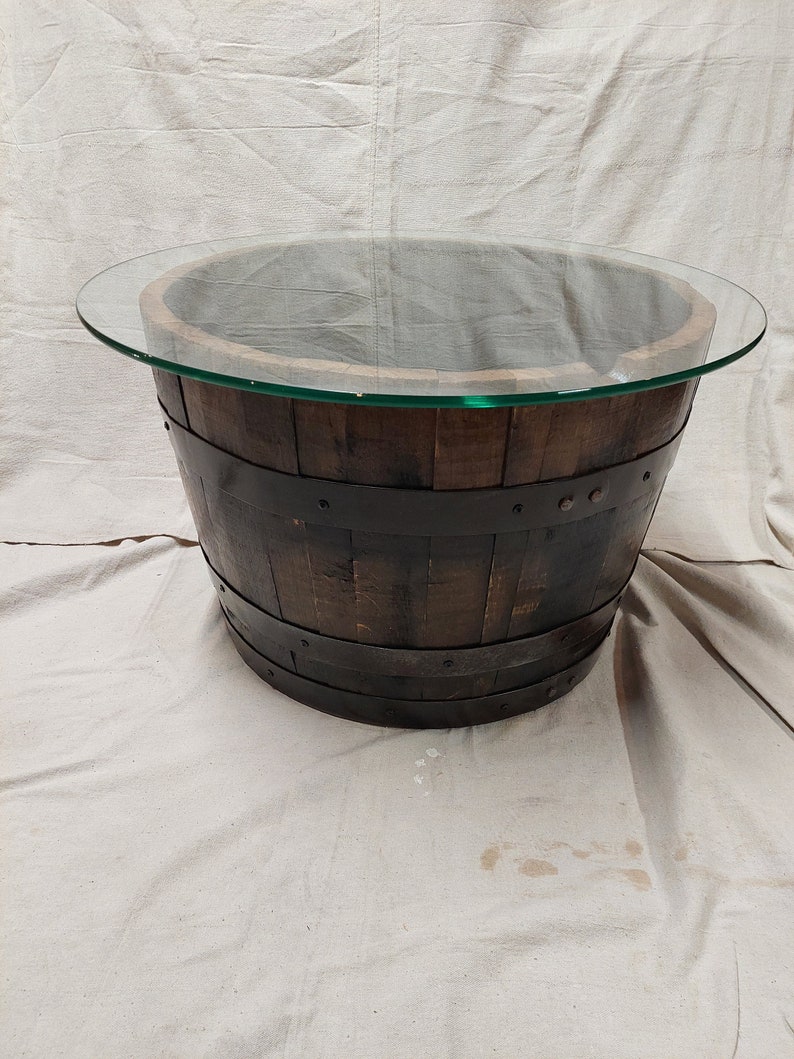 May include: A round coffee table made from a repurposed wooden barrel. The barrel is dark brown with black metal bands and a clear glass top. The table has a rustic, industrial design.