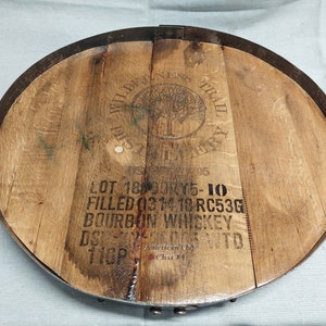 May include: A round wooden tray made from a reclaimed whiskey barrel with a metal rim. The tray has a brown wood grain and a black metal rim. The tray has a label that reads "WILDERNESS TRAIL DISTILLERY" and "LOT 18 DOK15-10 FILLED 0314 18 RC53G BOURBON WHISKEY DS  WTO 110P American Char 24".