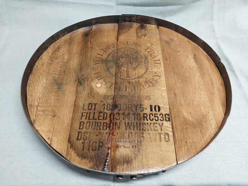 May include: A round wooden tray made from a reclaimed whiskey barrel with a metal rim. The tray has a brown wood grain and a black metal rim. The tray has a label that reads "WILDERNESS TRAIL DISTILLERY" and "LOT 18 DOK15-10 FILLED 0314 18 RC53G BOURBON WHISKEY DS  WTO 110P American Char 24".