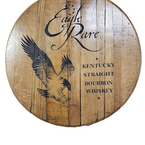 Pode incluir: Uma tampa de barril circular de madeira com um gr&aacute;fico preto de uma &aacute;guia voando e o texto Eagle Rare, Kentucky Straight Bourbon Whiskey. A madeira tem um gr&atilde;o castanho claro natural com t&aacute;buas verticais unidas numa forma redonda.