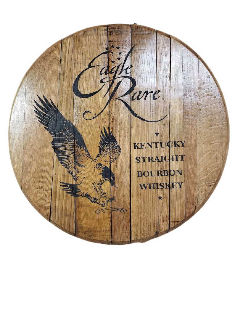 Pode incluir: Uma tampa de barril circular de madeira com um gr&aacute;fico preto de uma &aacute;guia voando e o texto Eagle Rare, Kentucky Straight Bourbon Whiskey. A madeira tem um gr&atilde;o castanho claro natural com t&aacute;buas verticais unidas numa forma redonda.