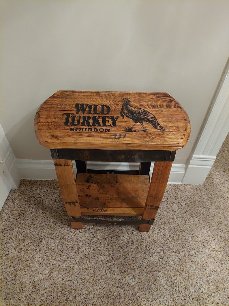May include: A wooden stool with a Wild Turkey Bourbon barrel top. The top is stained brown and features the Wild Turkey logo and a turkey illustration.