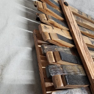 K&ouml;nnte beinhalten: Ein Wandregal aus Holz, hergestellt aus wiederverwendeten Weinfassdauben. Das Regal ist dunkelbraun gebeizt und hat ein rustikales Aussehen. Das Regal ist mit Schrauben an der Wand befestigt und verf&uuml;gt &uuml;ber eine schwarze Metallhalterung.