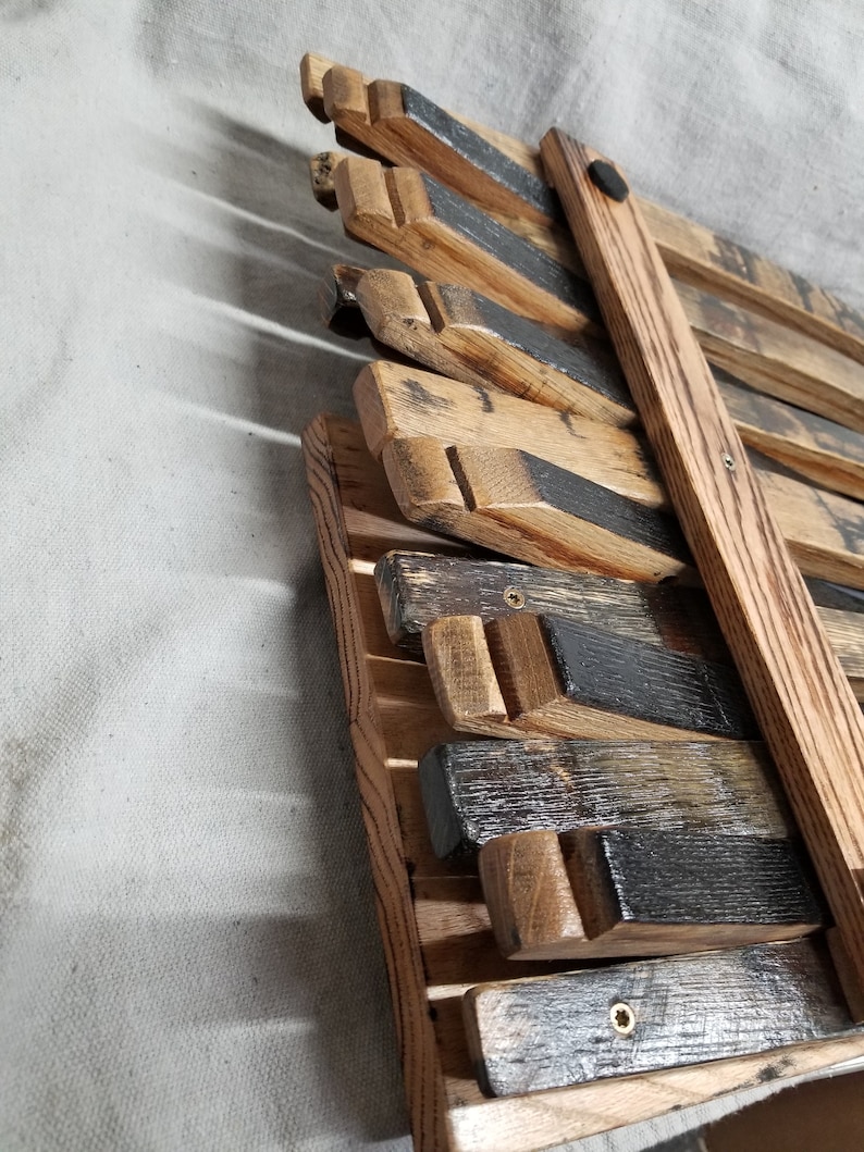 K&ouml;nnte beinhalten: Ein Wandregal aus Holz, hergestellt aus wiederverwendeten Weinfassdauben. Das Regal ist dunkelbraun gebeizt und hat ein rustikales Aussehen. Das Regal ist mit Schrauben an der Wand befestigt und verf&uuml;gt &uuml;ber eine schwarze Metallhalterung.