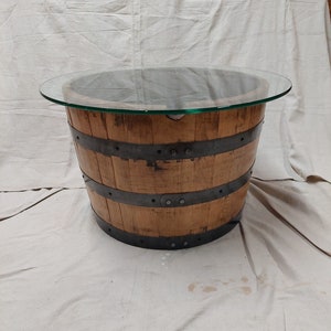 May include: A round coffee table made from a repurposed wooden barrel. The barrel has metal bands and a clear glass top. The wood has a natural finish, and the table is suitable for a living room. The table is approximately 76 cm in diameter.