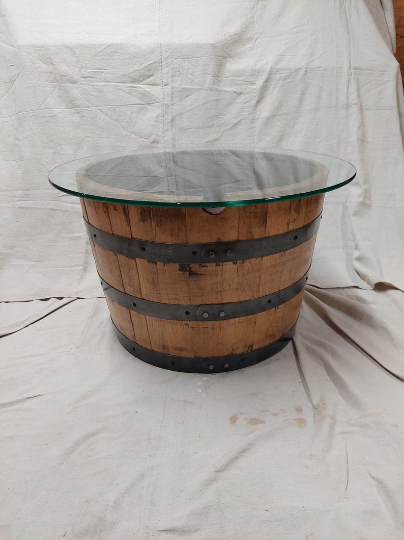 May include: A round coffee table made from a repurposed wooden barrel. The barrel has metal bands and a clear glass top. The wood has a natural finish, and the table is suitable for a living room. The table is approximately 76 cm in diameter.