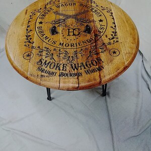 Pode incluir: Uma mesa redonda de madeira feita a partir de uma tampa de barril com pernas de metal preto. O topo apresenta uma impress&atilde;o gr&aacute;fica preta de dois rev&oacute;lveres cruzados, o texto "SMOKE WAGON BOURBON", "BIBAMUS MORIENDUM EST" e "SMOKE WAGON STRAIGHT BOURBON WHISKEY".