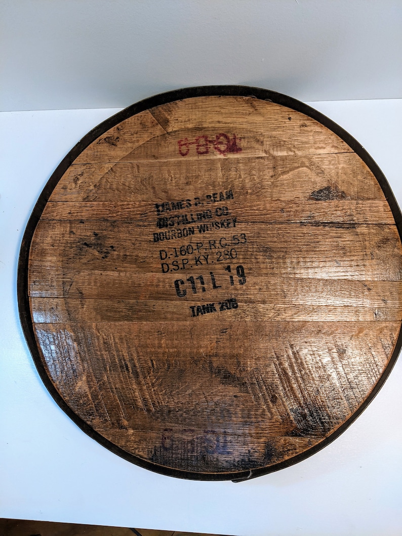 May include: A wooden barrel head with a black metal rim. The barrel head is marked with the text "JAMES B. BEAM DISTILLING CO. BOURBON WHISKEY D.-160-P.R.C. 53 D.S.P. KY. 280 C11L 19 TANK 208".