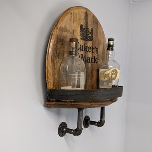 Op de afbeelding: Een houten vatenplank met zwarte metalen accenten en twee glazen flessen. De flessen zijn gelabeld "Woodford Reserve" en "Buffalo Trace" en het vat is gebrandmerkt "Maker's Mark".