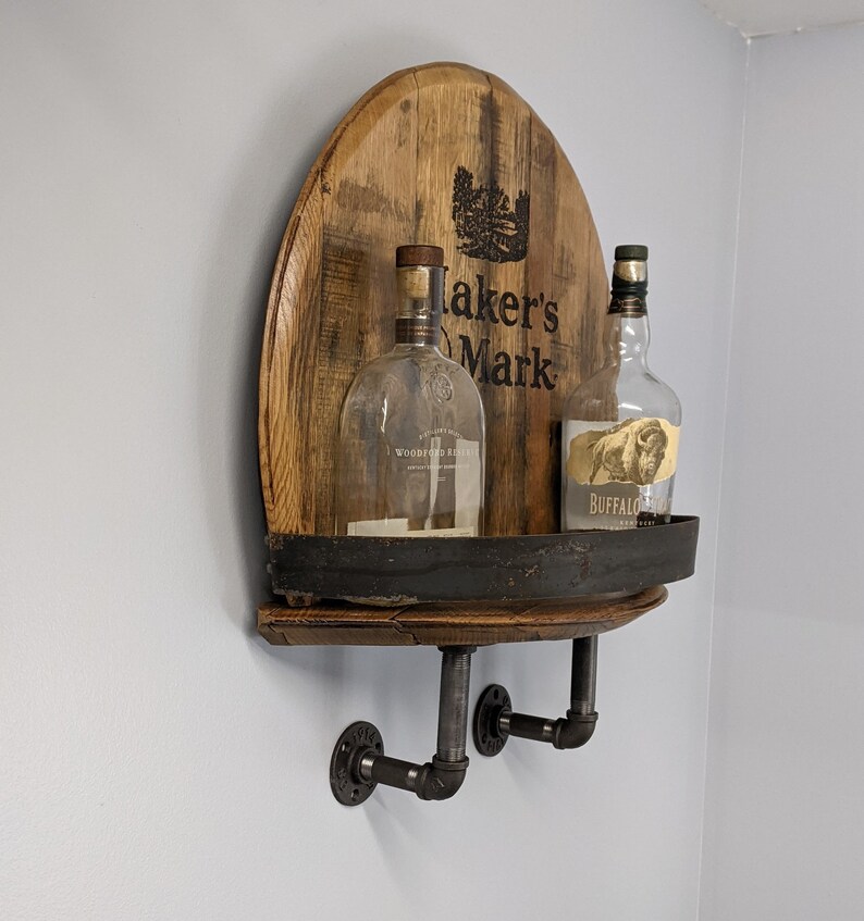 Op de afbeelding: Een houten vatenplank met zwarte metalen accenten en twee glazen flessen. De flessen zijn gelabeld "Woodford Reserve" en "Buffalo Trace" en het vat is gebrandmerkt "Maker's Mark".