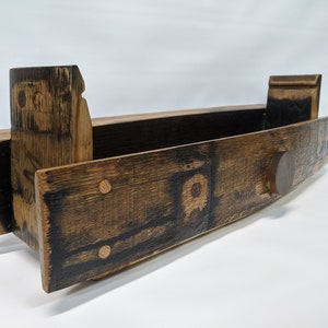 May include: A wooden shelf made from a repurposed barrel with a dark brown stain. The shelf has a rounded front edge and a small wooden knob on the right side. The shelf is made of wood with a natural grain pattern.