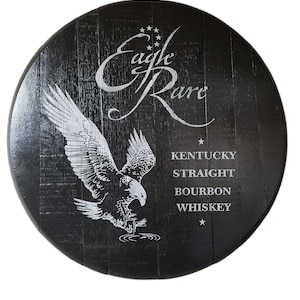 Pode incluir: Uma tampa de barril de madeira preta circular com um gr&aacute;fico branco de uma &aacute;guia em voo e o texto Eagle Rare Kentucky Straight Bourbon Whiskey. O veio da madeira &eacute; vis&iacute;vel sob o acabamento escuro, com estrelas brancas acima da marca.