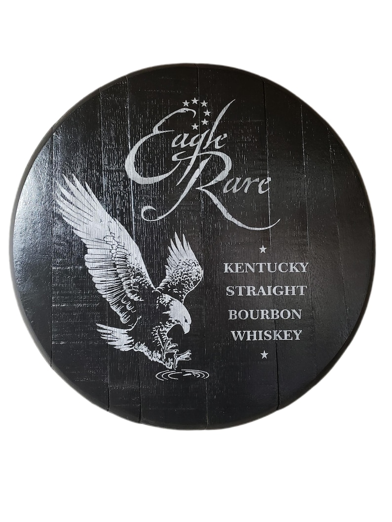 Pode incluir: Uma tampa de barril de madeira preta circular com um gr&aacute;fico branco de uma &aacute;guia em voo e o texto Eagle Rare Kentucky Straight Bourbon Whiskey. O veio da madeira &eacute; vis&iacute;vel sob o acabamento escuro, com estrelas brancas acima da marca.