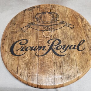 Pode incluir: Uma tampa de barril de madeira circular com um acabamento de gr&atilde;o de madeira castanho claro natural. A superf&iacute;cie apresenta o log&oacute;tipo Crown Royal impresso a preto com um gr&aacute;fico de coroa detalhado acima do texto, mostrando a textura da madeira.