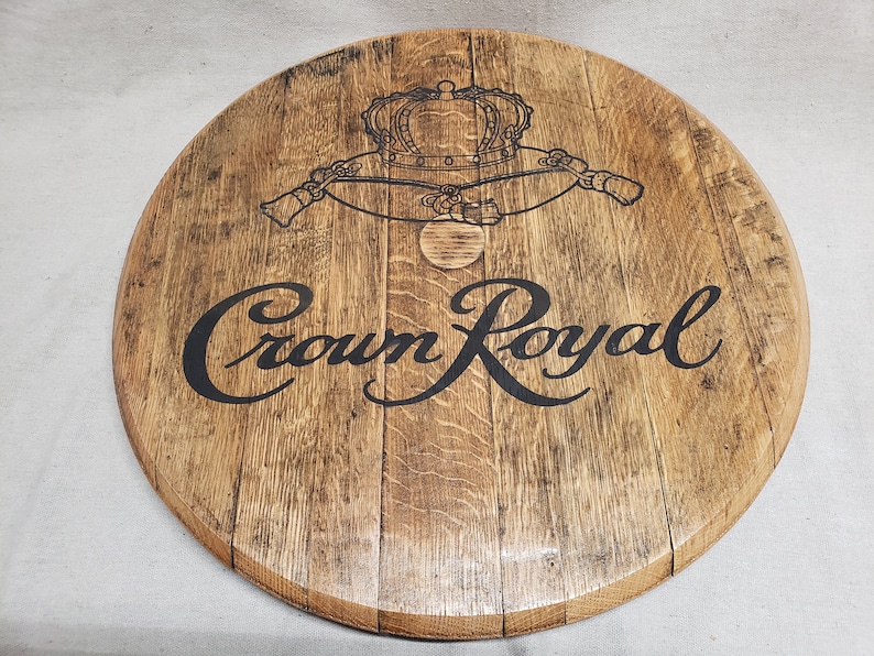 Pode incluir: Uma tampa de barril de madeira circular com um acabamento de gr&atilde;o de madeira castanho claro natural. A superf&iacute;cie apresenta o log&oacute;tipo Crown Royal impresso a preto com um gr&aacute;fico de coroa detalhado acima do texto, mostrando a textura da madeira.