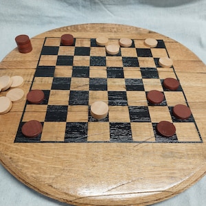 Pode incluir: Um tabuleiro de madeira circular com um padr&atilde;o de xadrez quadrado com quadrados pretos e de madeira natural alternados. O tabuleiro &eacute; acompanhado por v&aacute;rias pe&ccedil;as de jogo redondas em castanho escuro e bege claro, algumas colocadas no tabuleiro.