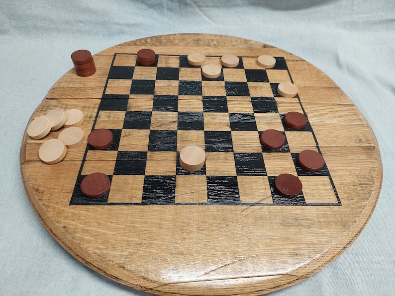 Pode incluir: Um tabuleiro de madeira circular com um padr&atilde;o de xadrez quadrado com quadrados pretos e de madeira natural alternados. O tabuleiro &eacute; acompanhado por v&aacute;rias pe&ccedil;as de jogo redondas em castanho escuro e bege claro, algumas colocadas no tabuleiro.