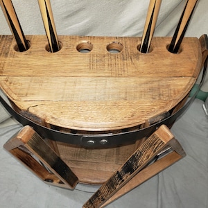 May include: A semi-circular wooden pool cue rack crafted from barrel staves with a natural oak finish. The rack features six circular cutouts along the top edge for holding cues, secured by a dark metal band with two visible rivets at the front.