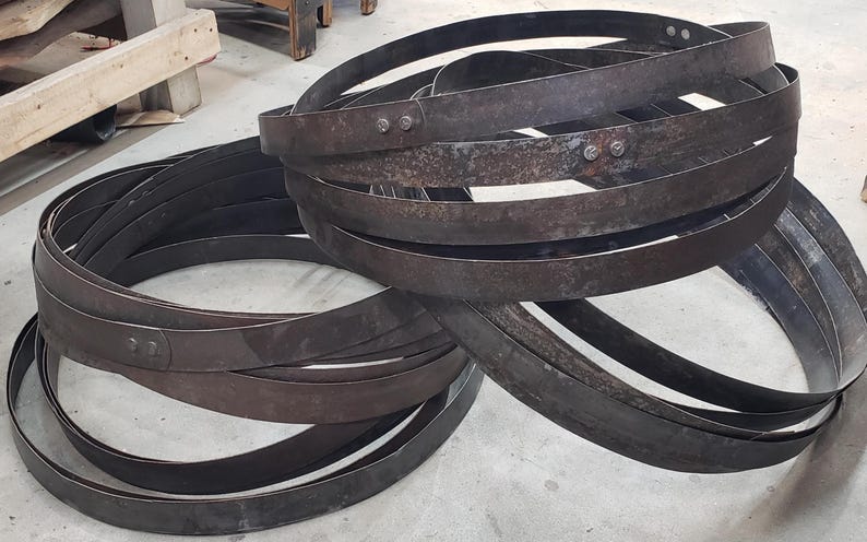 Pode incluir: Uma cole&ccedil;&atilde;o de aros de barril de metal marrom escuro, alguns com rebites, s&atilde;o empilhados e dispostos sobre uma superf&iacute;cie cinza claro. Os aros s&atilde;o curvos e parecem ser feitos de metal desgastado.