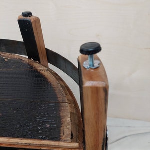 K&ouml;nnte beinhalten: Eine Struktur aus Fassdauben mit einer dunkel verkohlten Innenfl&auml;che und einer nat&uuml;rlich hellbraunen Au&szlig;enseite. Das Objekt umfasst ein gebogenes Metallband und zwei verstellbare, schwarze Kunststoff-Stellf&uuml;&szlig;e mit Gewindestangen.