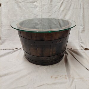 May include: A round coffee table made from a repurposed wooden barrel. The barrel is dark brown with metal bands and a clear glass top. The table is approximately 76 cm in diameter.