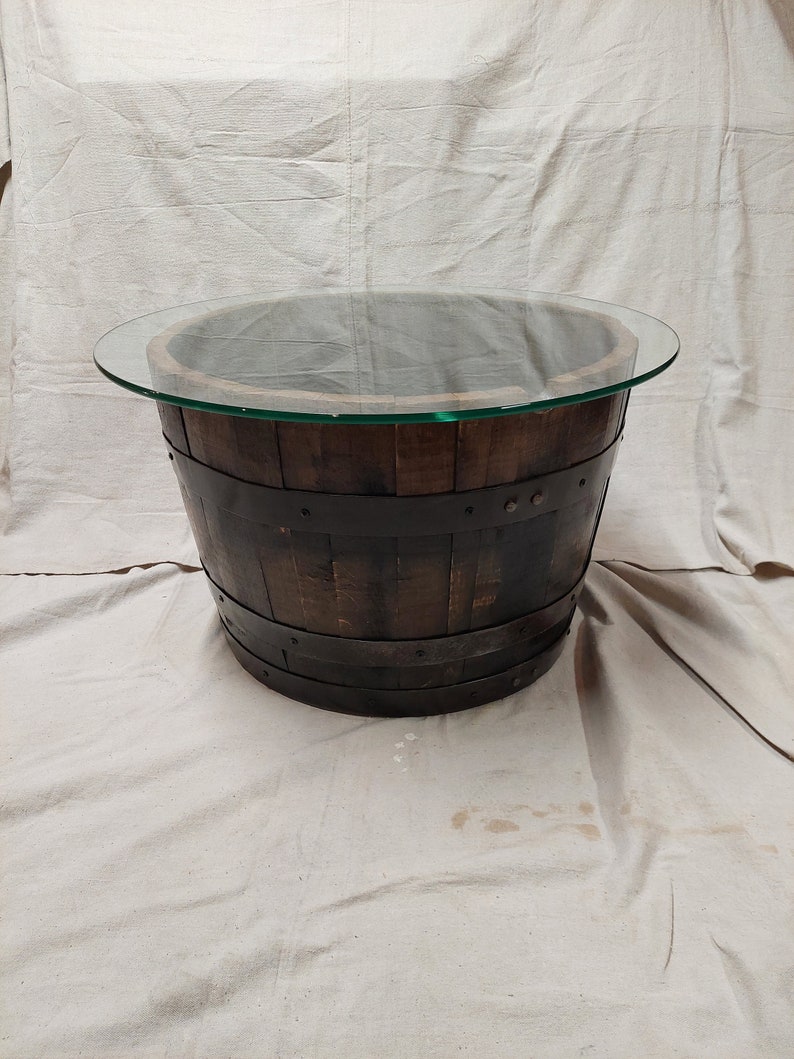 May include: A round coffee table made from a repurposed wooden barrel. The barrel is dark brown with metal bands and a clear glass top. The table is approximately 76 cm in diameter.