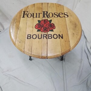 Pode incluir: Uma mesa redonda de madeira feita com aduelas de barril, apresentando o log&oacute;tipo Four Roses Bourbon em letras pretas com um gr&aacute;fico central de quatro rosas vermelhas. A superf&iacute;cie tem um acabamento polido e pernas de metal preto.