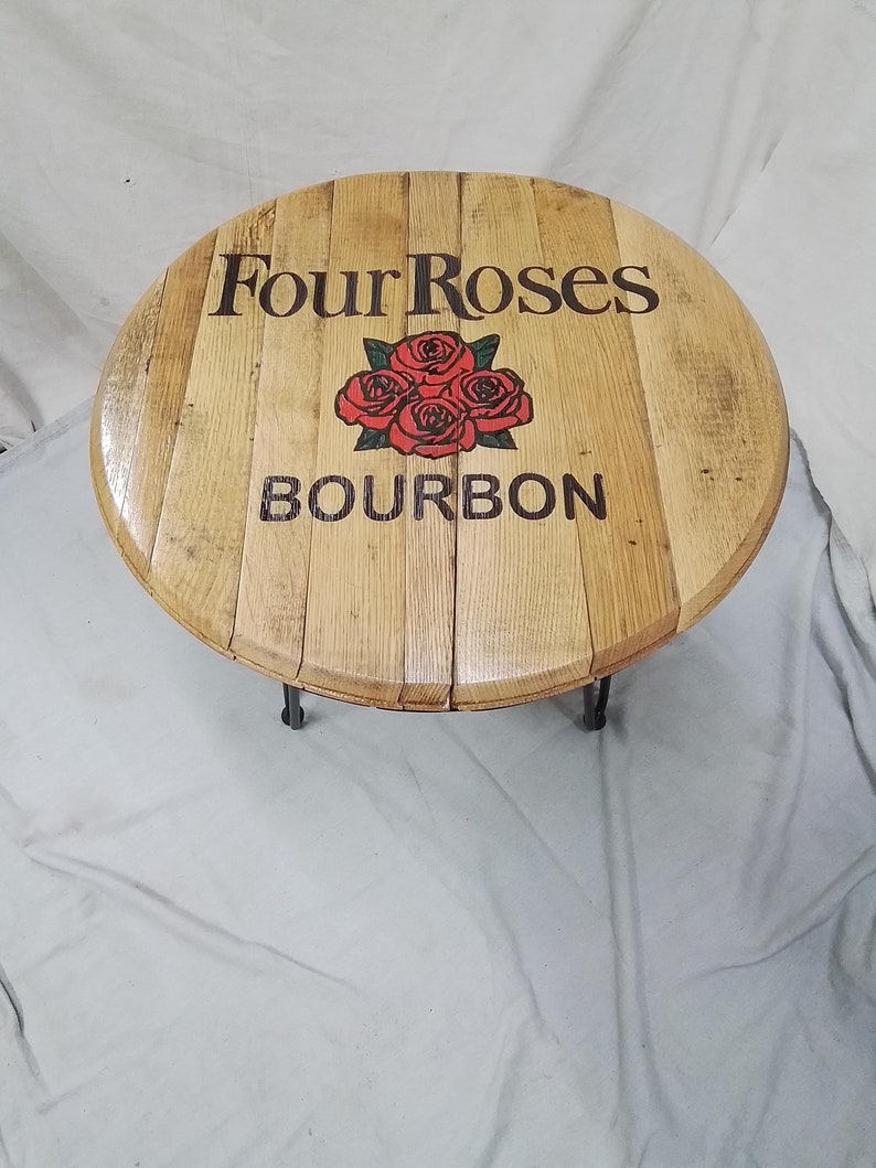 Pode incluir: Uma mesa redonda de madeira feita com aduelas de barril, apresentando o log&oacute;tipo Four Roses Bourbon em letras pretas com um gr&aacute;fico central de quatro rosas vermelhas. A superf&iacute;cie tem um acabamento polido e pernas de metal preto.