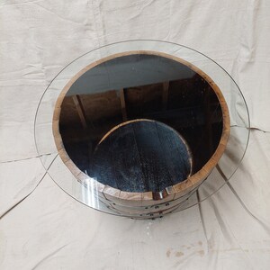May include: A circular coffee table constructed from a reclaimed wooden barrel. The barrel features metal bands and a transparent glass top. The interior of the barrel is dark, and the table rests on a neutral-toned surface.