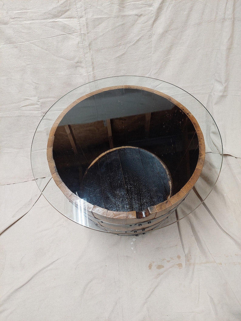 May include: A circular coffee table constructed from a reclaimed wooden barrel. The barrel features metal bands and a transparent glass top. The interior of the barrel is dark, and the table rests on a neutral-toned surface.