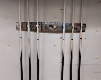 May include: A wooden pool cue rack with six cues. The rack is made of wood and has a curved design. The cues are black and white with a light-colored tip. The rack is mounted on a light-colored wooden wall.