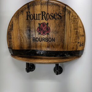 Op de afbeelding: Een houten plank gemaakt van een gerecycled Four Roses Bourbon-vat met zwarte metalen pijpbeugels. De plank is gemonteerd aan een witte muur.