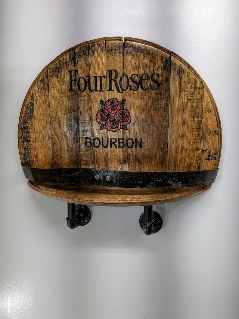 Op de afbeelding: Een houten plank gemaakt van een gerecycled Four Roses Bourbon-vat met zwarte metalen pijpbeugels. De plank is gemonteerd aan een witte muur.