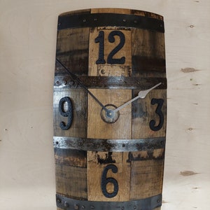 May include: A rustic wooden clock made from a repurposed barrel with metal bands and black Roman numeral hour markers. The clock face is made of wood and has a silver-colored hour and minute hand.