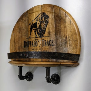 Op de afbeelding: Een houten wandplank met een Buffalo Trace Kentucky Straight Bourbon whiskey vaten bovenkant. De plank heeft twee zwarte metalen pijpbeugels. De vaten bovenkant heeft een zwart buffel silhouet en de tekst "Buffalo Trace Kentucky Straight Bourbon".