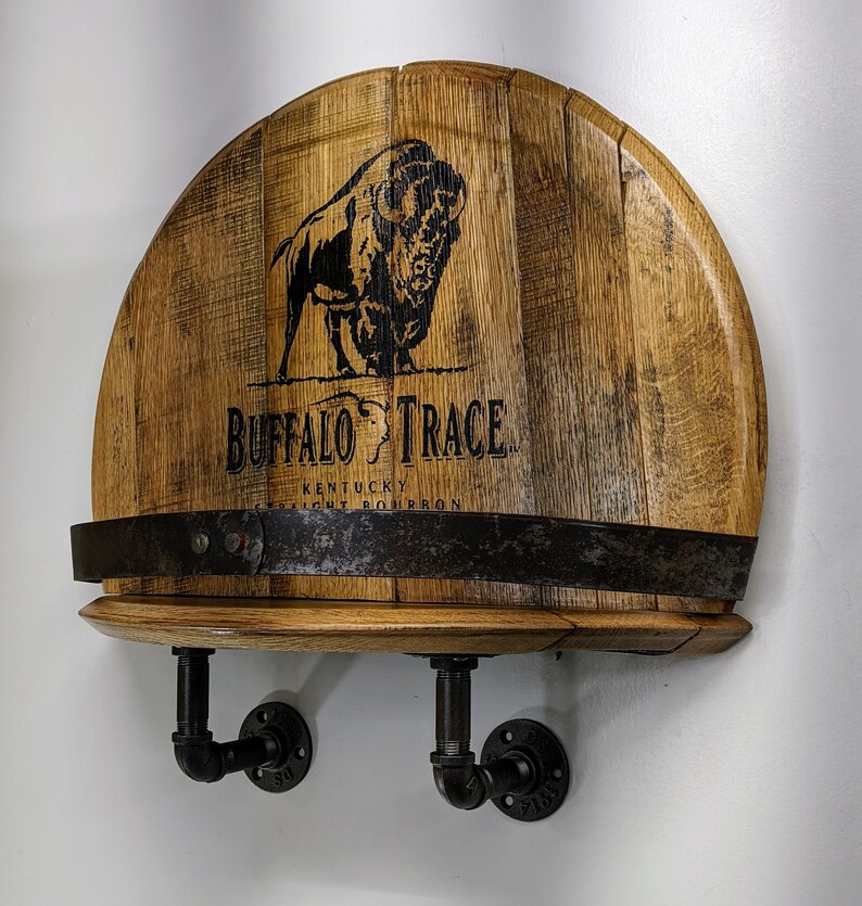 Op de afbeelding: Een houten wandplank met een Buffalo Trace Kentucky Straight Bourbon whiskey vaten bovenkant. De plank heeft twee zwarte metalen pijpbeugels. De vaten bovenkant heeft een zwart buffel silhouet en de tekst "Buffalo Trace Kentucky Straight Bourbon".