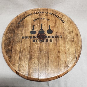 Op de afbeelding: Een ronde houten vatenkop met de tekst "Woodford Reserve Distillery" en "DSP-KY-52" erop gedrukt. De tekst "Bourbon Whiskey 1 RC 52 8 G" is ook op de vatenkop gedrukt, samen met drie gestileerde afbeeldingen van decanters.
