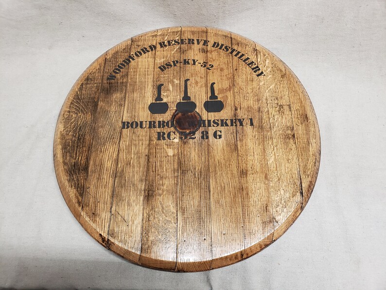 Op de afbeelding: Een ronde houten vatenkop met de tekst "Woodford Reserve Distillery" en "DSP-KY-52" erop gedrukt. De tekst "Bourbon Whiskey 1 RC 52 8 G" is ook op de vatenkop gedrukt, samen met drie gestileerde afbeeldingen van decanters.
