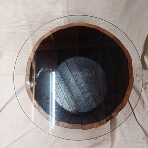 May include: Circular coffee table with a clear glass top. The base is a repurposed wooden barrel with a dark brown interior and a circular wooden base. The barrel's exterior is a light brown colour.