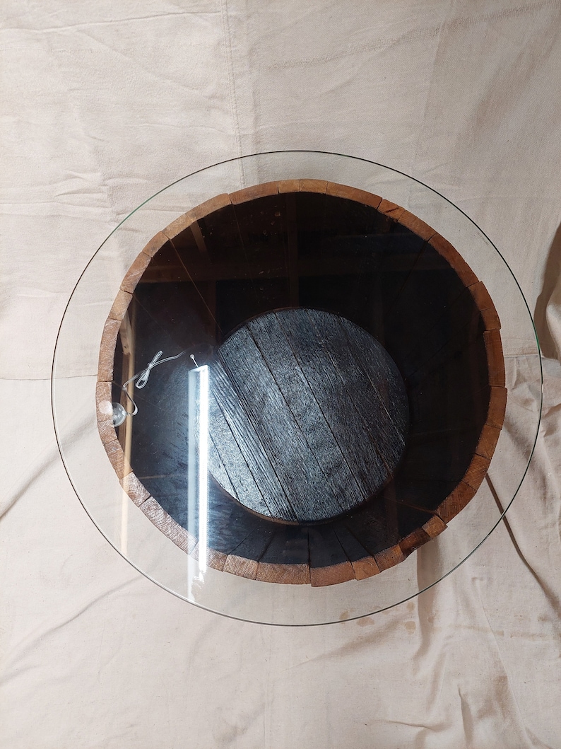 May include: Circular coffee table with a clear glass top. The base is a repurposed wooden barrel with a dark brown interior and a circular wooden base. The barrel's exterior is a light brown colour.