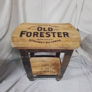May include: A wooden side table with a top made from a reclaimed bourbon barrel. The top is branded with the words "First Bottled Bourbon", "Est. 1870", "Old Forester", "Kentucky", and "Straight Bourbon".
