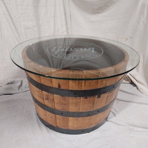 May include: A round coffee table made from a repurposed wooden barrel. The barrel is light brown with dark metal bands. A clear glass top covers the barrel, with the words "Gibson Guitars" etched into the glass.