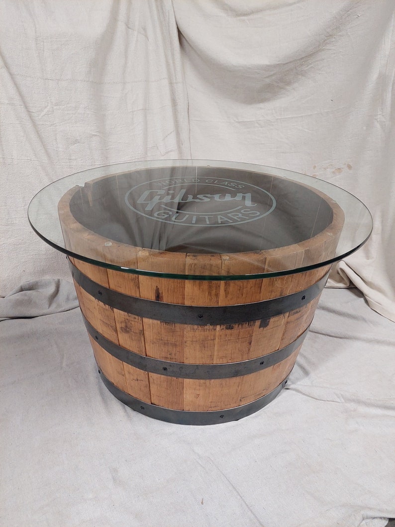 May include: A round coffee table made from a repurposed wooden barrel. The barrel is light brown with dark metal bands. A clear glass top covers the barrel, with the words "Gibson Guitars" etched into the glass.