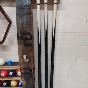 May include: A wooden wall-mounted rack for storing pool cues. The rack has a dark brown finish and features a barrel stave design. Four pool cues are stored in the rack, with black and white accents.