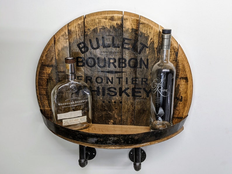 Op de afbeelding: Een houten vatenplank met een zwart metalen plank en twee doorzichtige glazen flessen. Het vat is gelabeld met "Bullet Bourbon Frontier Whiskey".